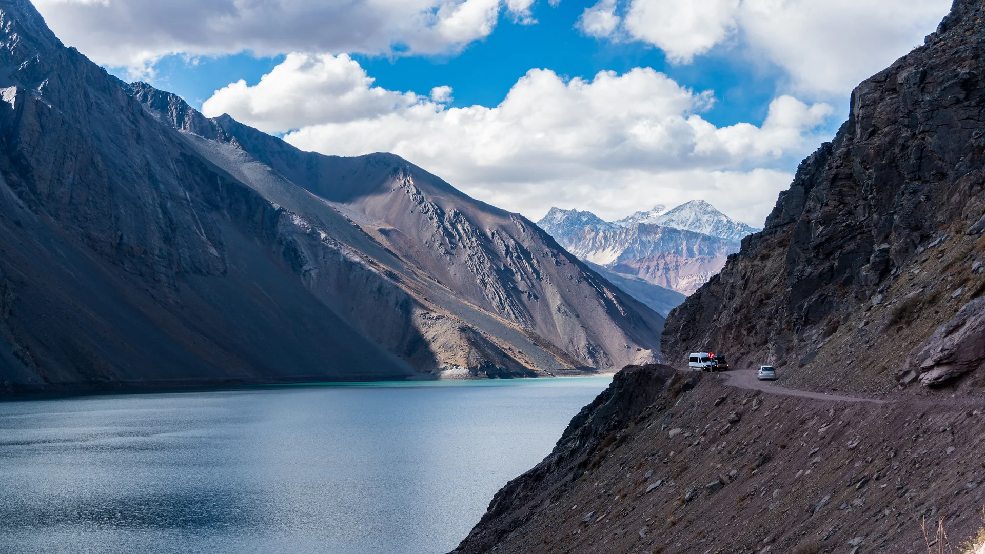 A van and a car on a narrow gravel switchback road above the turquoise Embalse El Yeso reservoir, framed by bare Andean peaks